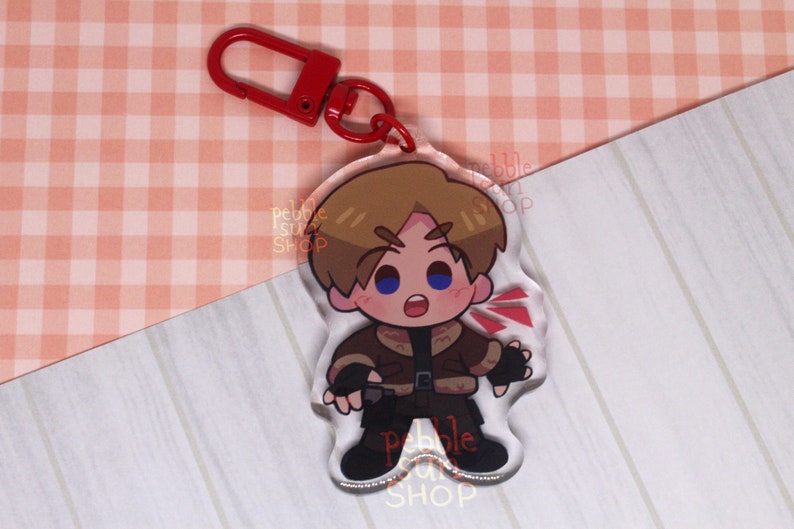 May include: A clear acrylic keychain featuring a cartoon character with brown hair, a blue jacket, and a red scarf. The character is holding a gun and has a surprised expression on their face. The keychain has a red metal ring.