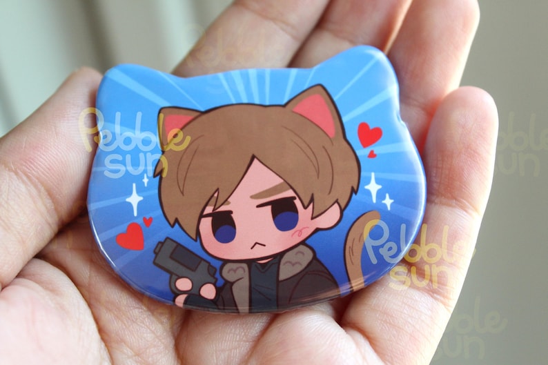 May include: A blue cat-shaped button featuring a cartoon character with brown hair, cat ears, and a serious expression, holding a gun. The design includes hearts and starbursts. The text "Pebble Sun" is visible.