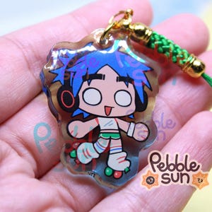 May include: A clear acrylic charm featuring a cartoon character with blue hair, headphones, and roller skates. The charm has a gold-colored clasp and a green cord. The charm is labeled with the text "Pebble Sun".