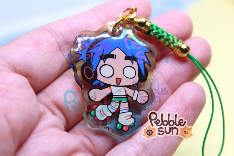 May include: A clear acrylic charm featuring a cartoon character with blue hair, headphones, and roller skates. The charm has a gold-colored clasp and a green cord. The charm is labeled with the text "Pebble Sun".