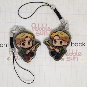 May include: A black phone strap with a clear charm featuring a cartoon character holding a weapon. The charm is attached to a black cord with a silver clasp. The text "Pebble Sun" is visible on the charm.