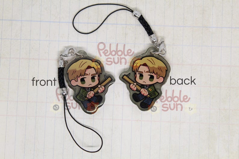 May include: A black phone strap with a clear charm featuring a cartoon character holding a weapon. The charm is attached to a black cord with a silver clasp. The text "Pebble Sun" is visible on the charm.