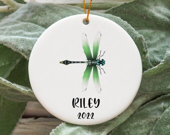 May include: White ceramic Christmas ornament with a green dragonfly illustration and the name "Riley" and the year "2022" printed in black.