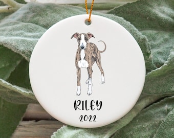May include: White ceramic Christmas ornament with a brown and white Greyhound dog illustration and the name "Riley" and the year "2022" in black text.