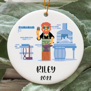 Pode incluir: Adorno de cerâmica branca com uma ilustração de desenho animado de um ferreiro trabalhando em uma bigorna. O ferreiro está vestindo uma camisa vermelha, jeans azul e um avental preto. O adorno tem o nome "Riley" e o ano "2022" impresso nele.