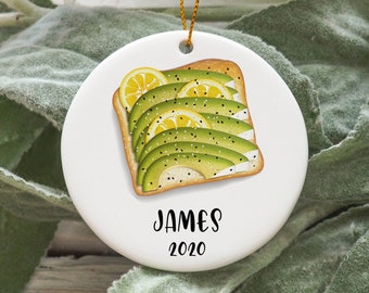 May include: White ceramic ornament with a cartoon illustration of avocado toast with lemon slices and black sesame seeds. The ornament is personalized with the name "JAMES" and the year "2020".