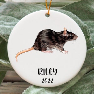 May include: A white ceramic bauble featuring a black and brown rat illustration, with the name "Riley" and the year "2022" in black text.