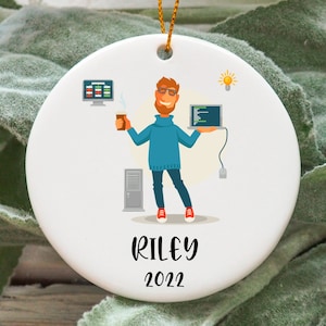 以下が含まれることがあります： コーヒーカップとラップトップを持った人の漫画イラストが描かれた白いセラミックオーナメント。オーナメントの下部に「RILEY 2022」の文字があります。