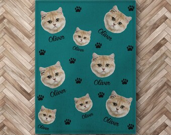 Könnte beinhalten: Eine tealblaue Fleece-Decke mit einem sich wiederholenden Muster aus einem Katzenkopf und dem Namen "Oliver" in Schwarz gedruckt. Das Katzen Gesicht ist hellbraun mit grünen Augen.