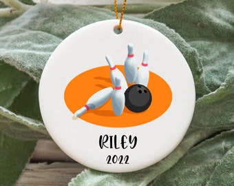 Puede incluir: Adorno navideño redondo de cerámica blanca con diseño de bolos. Muestra una bola de bolos negra y bolos blancos dentro de un óvalo naranja. El nombre "RILEY" y el año "2022" están impresos en negro.