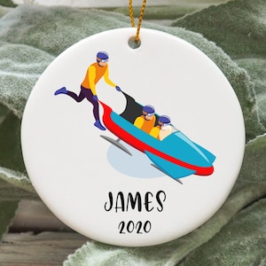 May include: A white ceramic ornament with a cartoon illustration of a bobsled team. The bobsled is blue, red, and white. A person in a yellow and blue outfit is pushing the bobsled. The ornament has the text "JAMES 2020" printed on it.