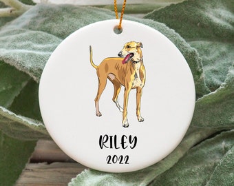 Op de afbeelding: Witte kerstbal van keramiek met een handgetekend ontwerp van een bruine en witte windhond. De kerstbal heeft de naam "Riley" en het jaar "2022" in zwart.