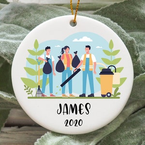 Peut inclure: Une décoration de Noël en céramique blanche avec une illustration de trois personnes ramassant des feuilles et des déchets. La décoration porte l'inscription "JAMES 2020" en bas.
