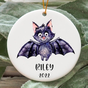 May include: A white ceramic bauble featuring a cartoon bat illustration and the text "Riley 2022".