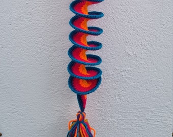 May include: A long, colorful crocheted spiral hanging from a white wall. The spiral is made of blue, pink, and orange yarn and has a tassel at the bottom.