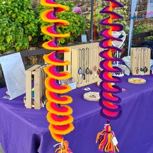May include: Two colorful crocheted spiral hanging decorations with tassels. The decorations are made with orange, yellow, pink, and purple yarn. They are hanging from a market stall.