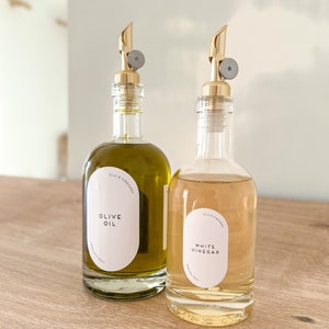K&ouml;nnte beinhalten: Zwei Glasflaschen mit goldenen Ausgie&szlig;ern und Etiketten. Die Flasche links ist mit Oliven&ouml;l gef&uuml;llt und tr&auml;gt ein Etikett mit der Aufschrift "Oils & Vinegars Olive Oil". Die Flasche rechts ist mit wei&szlig;em Essig gef&uuml;llt und tr&auml;gt ein Etikett mit der Aufschrift "Oils & Vinegars White Vinegar".
