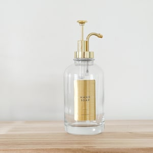 May include: Clear glass hand soap dispenser with a gold-colored pump and a gold label that reads "HAND SOAP". The dispenser is sitting on a light-colored wooden surface.