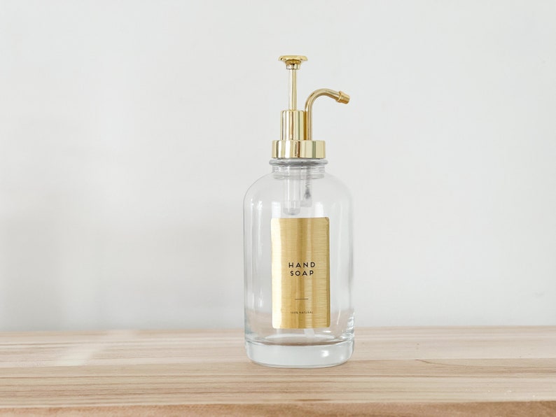 May include: A clear glass bottle with a gold pump dispenser and a gold label that reads "HAND SOAP". The bottle is sitting on a wooden surface.