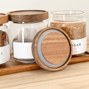 May include: Three glass jars with wooden lids, filled with brown sugar, tea bags, and coffee grounds. The jars are sitting on a wooden tray. The lid of one of the jars is off and sitting on the tray.