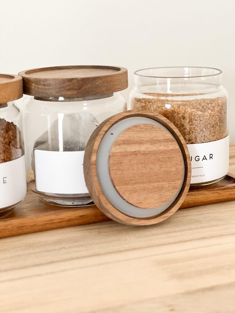 May include: Three glass jars with wooden lids, filled with brown sugar, tea bags, and coffee grounds. The jars are sitting on a wooden tray. The lid of one of the jars is off and sitting on the tray.