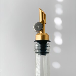 Puede incluir: Un vertedor de botella dorado con un tap&oacute;n de goma negro y un anillo plateado. El vertedor est&aacute; unido a una botella de vidrio transparente.