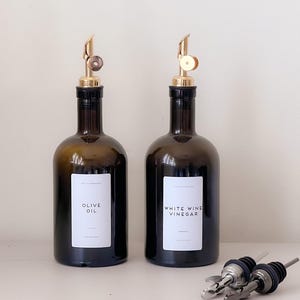 Puede incluir: Dos botellas de vidrio ámbar oscuro con vertedores de color dorado. Cada botella tiene una etiqueta rectangular blanca. Una etiqueta dice OLIVE OIL y la otra WHITE WINE VINEGAR. Dos vertedores de metal plateado adicionales se encuentran cerca.