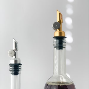 Puede incluir: Dos botellas de vidrio con boquillas de vertido. La botella de la izquierda es transparente y est&aacute; vac&iacute;a. La botella de la derecha est&aacute; llena de un l&iacute;quido oscuro y tiene una boquilla dorada.