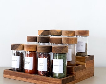 Peut inclure: Un porte-épices en bois avec un design à plusieurs niveaux contient des bocaux en verre avec des couvercles en bois. Les bocaux sont remplis de différentes épices, notamment des clous de girofle, du paprika fumé, des flocons de piment et du persil. Les étiquettes sur les bocaux sont blanches avec du texte noir.