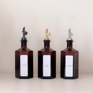 May include: Three amber glass bottles with metal pour spouts. Each bottle has a white label with black text: "VINEGAR", "OLIVE OIL", and "SUNFLOWER OIL". The bottles are hexagonal and sit on a white surface.