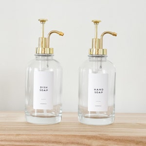 May include: Two clear glass soap dispensers with gold pumps and labels that read "Dish Soap" and "Hand Soap".
