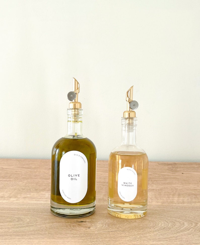 K&ouml;nnte beinhalten: Zwei Glasflaschen mit goldenen Ausgie&szlig;ern und Etiketten. Die Flasche links ist mit Oliven&ouml;l gef&uuml;llt und mit "Oliven&ouml;l" beschriftet. Die Flasche rechts ist mit Wei&szlig;weinessig gef&uuml;llt und mit "Wei&szlig;weinessig" beschriftet.