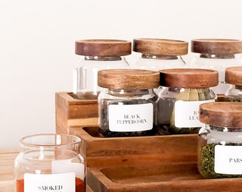 Könnte beinhalten: Ein Gewürzregal aus Holz mit Glasbehältern, die mit Gewürzen gefüllt sind. Die Behälter sind mit den Namen der Gewürze beschriftet, darunter "Geräucherter Paprika", "Schwarzer Pfefferkorn", "Lorbeerblatt" und "Petersilie".