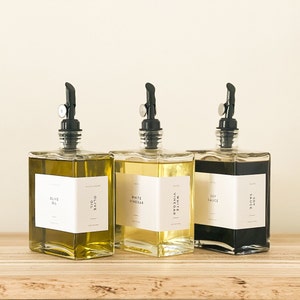 May include: Three glass bottles with black pump tops and white labels. The bottles contain olive oil, white vinegar, and soy sauce. The bottles are sitting on a wooden surface.