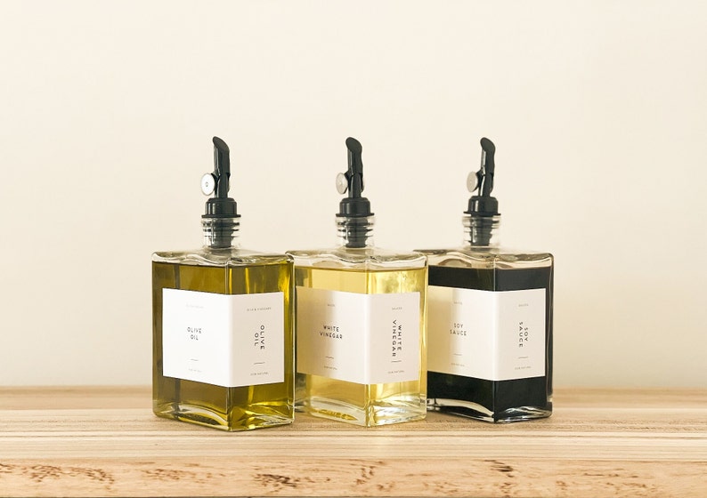 May include: Three glass bottles with black pump tops and white labels. The bottles contain olive oil, white vinegar, and soy sauce. The bottles are sitting on a wooden surface.