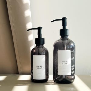 May include: Two dark grey glass soap dispensers with black pumps. The smaller bottle is labelled "HAND SOAP", and the larger one is labelled "DISH SOAP". Both have white labels and are on a light-coloured surface.