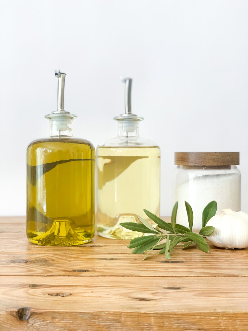 May include: Two glass bottles with pour spouts filled with olive oil and vinegar, a jar of salt, a clove of garlic, and a sprig of green herbs on a wooden table.