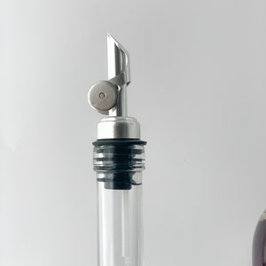 Puede incluir: Un vertedor de botella plateado con un sello de goma negro y una botella de vidrio transparente.