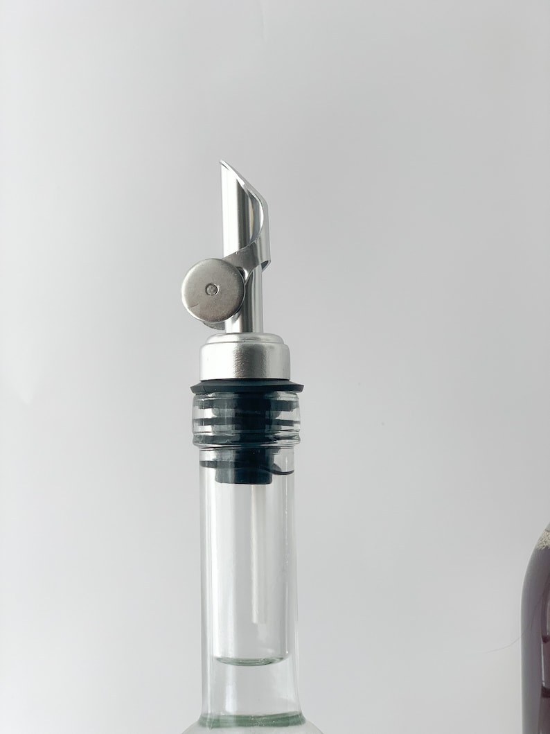 Puede incluir: Un vertedor de botella plateado con un sello de goma negro y una botella de vidrio transparente.