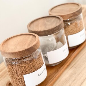 May include: Three glass jars with wooden lids and labels on a wooden tray. The jars contain coffee, tea, and sugar. The labels are white with black text. The text on the labels reads "Coffee", "Tea", and "Sugar".