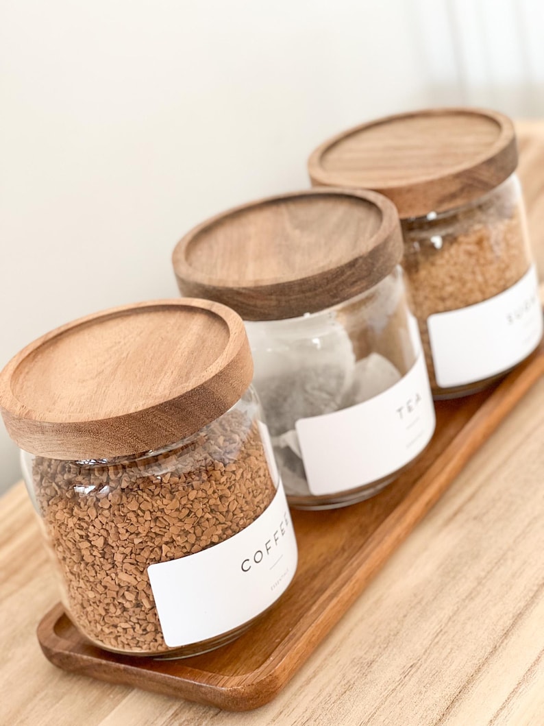 May include: Three glass jars with wooden lids and labels on a wooden tray. The jars contain coffee, tea, and sugar. The labels are white with black text. The text on the labels reads "Coffee", "Tea", and "Sugar".