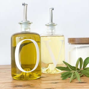 May include: Two glass bottles with clear labels and white lettering, one labeled "O" and the other labeled "V", filled with olive oil and vinegar, respectively. The bottles are on a wooden surface with a sprig of green leaves.
