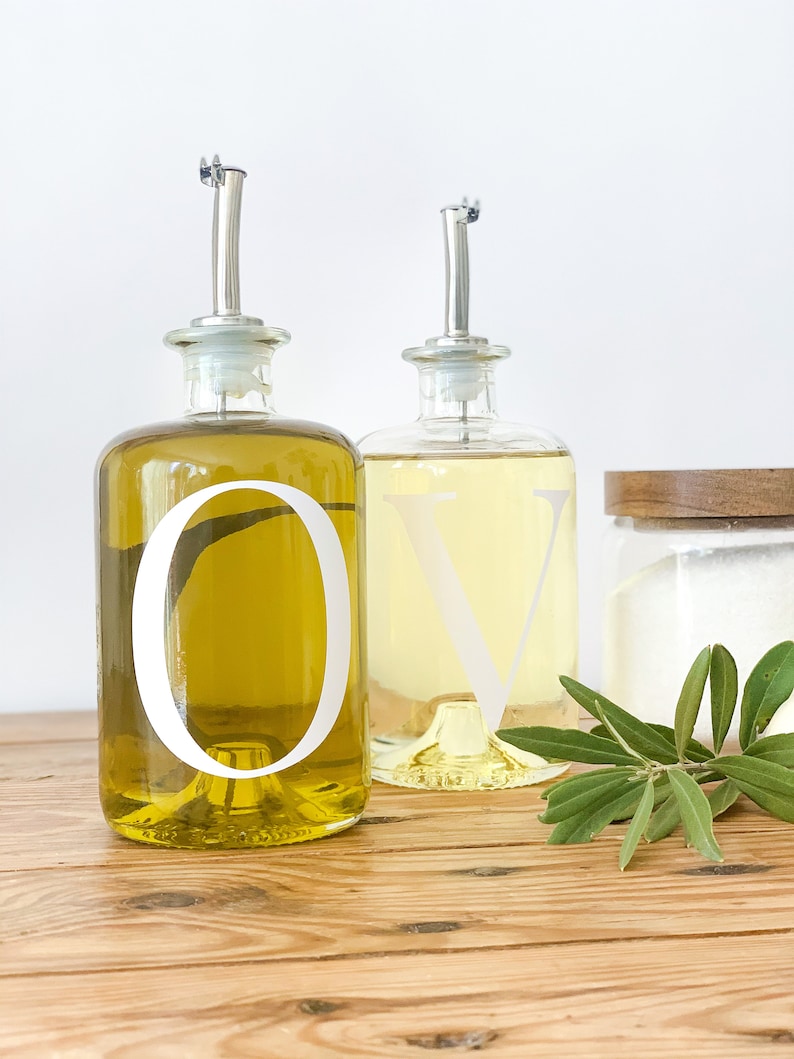 May include: Two glass bottles with clear labels and white lettering, one labeled "O" and the other labeled "V", filled with olive oil and vinegar, respectively. The bottles are on a wooden surface with a sprig of green leaves.