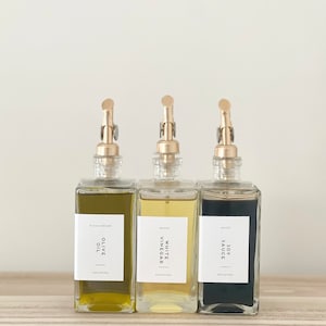 May include: Three glass bottles with gold pump tops and labels. The bottles contain olive oil, white vinegar, and soy sauce. The labels are white with black text and say "Olive Oil", "White Vinegar", and "Soy Sauce".