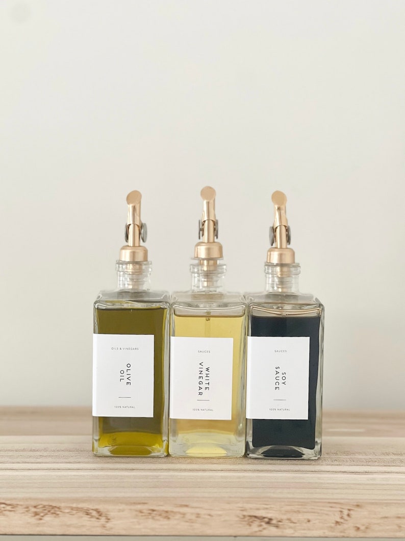 May include: Three glass bottles with gold pump tops and labels. The bottles contain olive oil, white vinegar, and soy sauce. The labels are white with black text and say "Olive Oil", "White Vinegar", and "Soy Sauce".