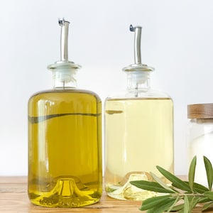 May include: Two clear glass bottles with pour spouts filled with olive oil and vinegar, sitting on a wooden surface with a sprig of green herbs.