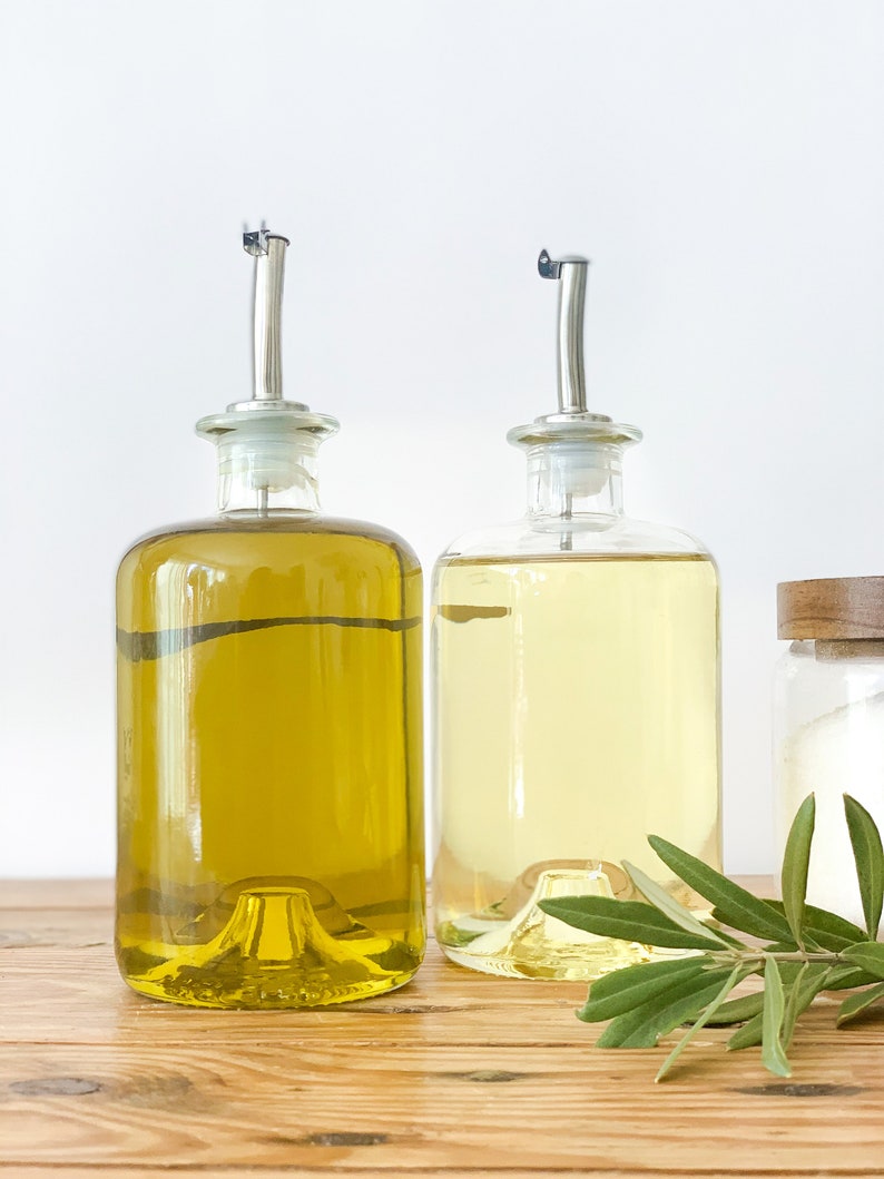 May include: Two clear glass bottles with pour spouts filled with olive oil and vinegar, sitting on a wooden surface with a sprig of green herbs.
