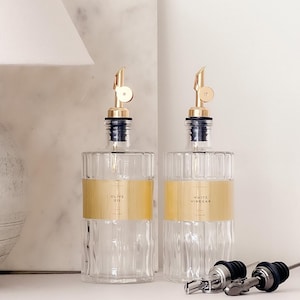 Può includere: Due bottiglie in vetro trasparente con scanalature verticali e beccucci versatori in metallo dorato. Ogni bottiglia presenta un'ampia etichetta orizzontale dorata. Un'etichetta riporta "OLIVE OIL" e l'altra "WHITE VINEGAR". Due beccucci extra vicini.