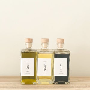 May include: Three glass bottles with cork tops and labels. The bottles contain olive oil, white vinegar, and soy sauce. The labels are white with black text and say "Olive Oil", "White Vinegar", and "Soy Sauce".