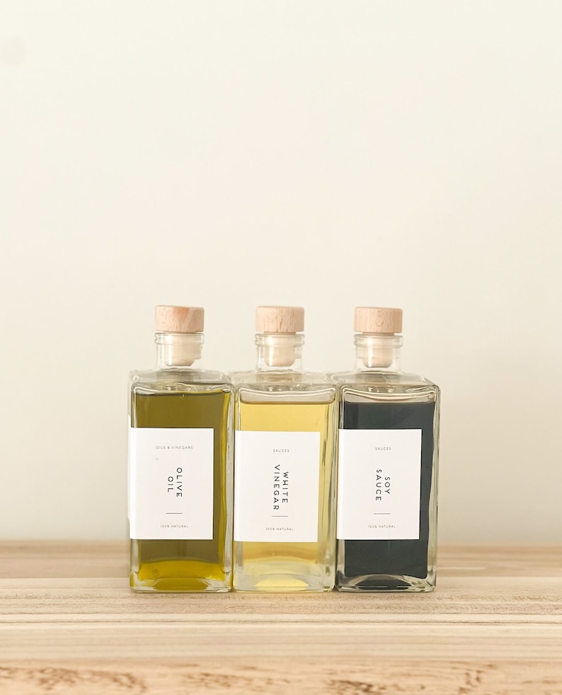 May include: Three glass bottles with cork tops and labels. The bottles contain olive oil, white vinegar, and soy sauce. The labels are white with black text and say "Olive Oil", "White Vinegar", and "Soy Sauce".